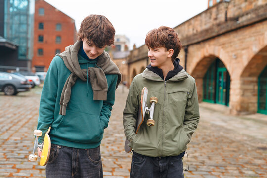 Two boys teenagers walking in a city area while holding skateboards and talking to each other