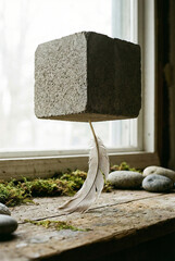 A large stone cube suspended in mid-air by a delicate feather near a window with stones and moss on the wooden sill