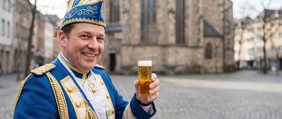 Smiling man in traditional blue carnival guard uniform holding a glass of beer. German street festival celebration in Cologne with blurred church background.