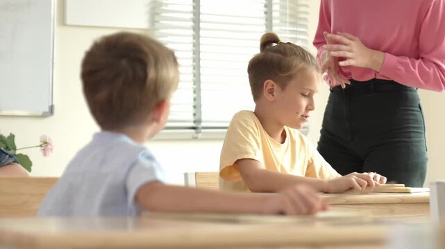 Beautiful teacher explaining test while caucasian boy doing classwork and listening explanation. Cute child looking at teacher while taking a notes or writing answer in paper at classroom. Pedagogy.