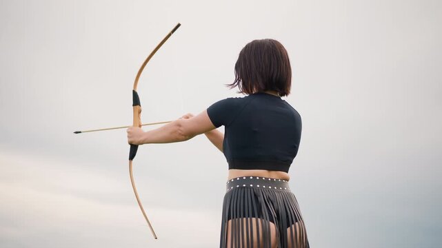 woman archer poised with bow facing horizon, leather fringe skirt and cropped top, drawing arrow with steady stance under overcast sky, cinematic slow motion mood, solitary warrior ritual, focused