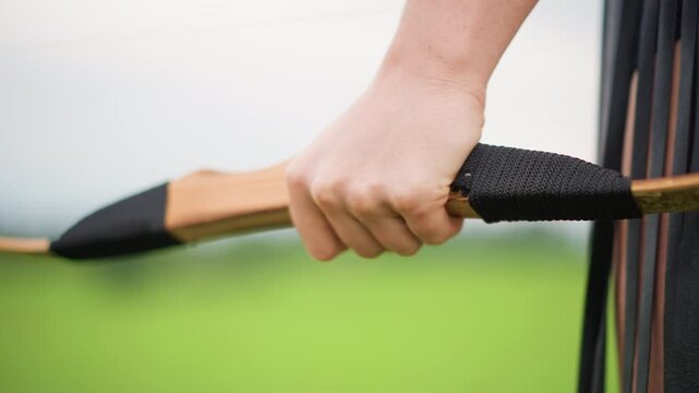 closeup caucasian female hand gripping bow, leatherwrapped handle, green field backdrop, steady practiced grip, subtle tension in fingers, solitary warrior atmosphere, warm wood tones, detailed