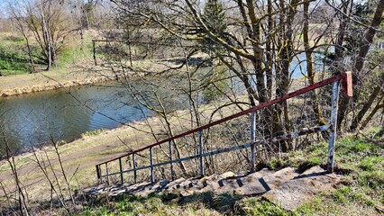 Fototapeta premium The river flows between grassy banks with dirt roads. Birch, willow and spruce trees grow nearby. A pipeline is installed above the river. A concrete staircase with metal railings leads to the water