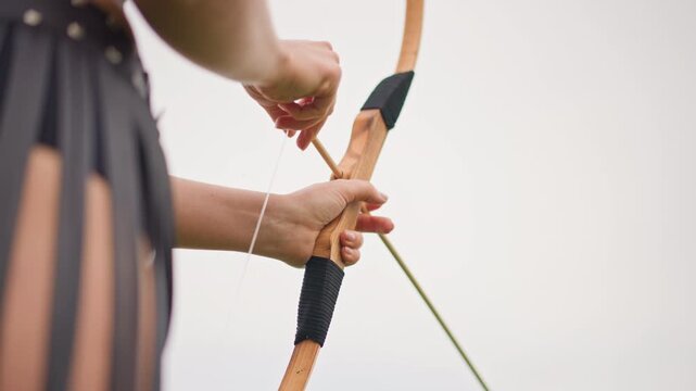 Closeup archer hands nocking arrow, leather fringe skirt and studded belt visible, wooden bow gripped tight, bowstring tension building against overcast sky, focused training moment with precise