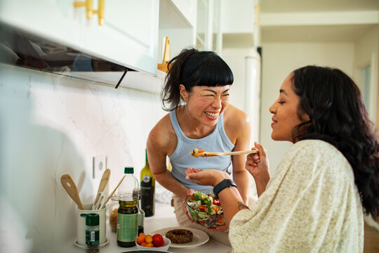 Two women tasting fresh salad in home kitchen
