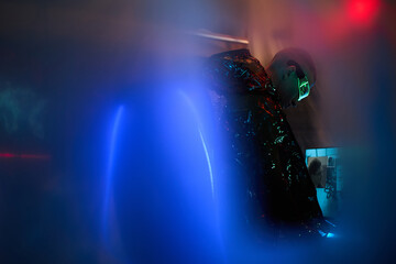 Young adult Caucasian man wearing reflective jacket and neon glasses standing in dark room using computer equipment, illuminated by blue and red lights, engaging in hacking activity