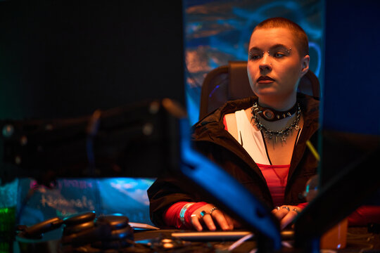 Young adult Caucasian woman sitting at computer desk typing on keyboard, wearing spiked choker and facial piercings, concentrating on multiple monitors in dark room