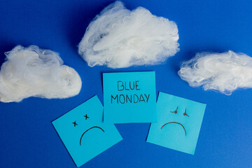 Creative flat lay composition on blue background with cotton clouds and sticky notes featuring sad faces and Blue Monday text.
