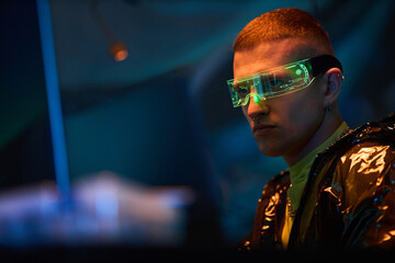 Young adult Caucasian man wearing futuristic augmented reality glasses sitting at computer workstation hacking digital system in dark room with neon lighting