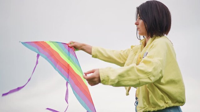 young woman launching rainbow kite, yellow jacket testing wind and unfurling colorful fabric outstretched arm holds kite while purple ribbons flutter against overcast sky, casual jeans and relaxed
