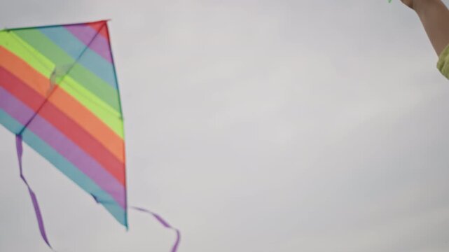 closeup colorful kite wing against sky, vivid rainbow panels and purple tails fluttering, hand adjusting string, overcast light and soft cloud backdrop, fabric texture and stitched seams visible,