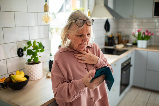 Concerned senior woman reading smartphone in home kitchen