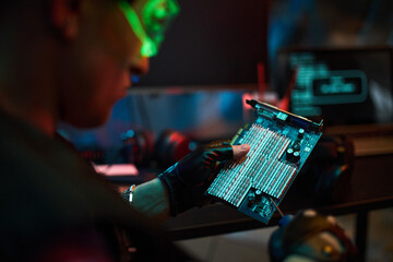 Young man wearing gloves holding computer circuit board examining hardware with blurred laptop displaying code in background suggesting cybersecurity or hacking activity