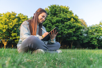 Young adult woman sitting on grass, writing in a notebook with trees in the background