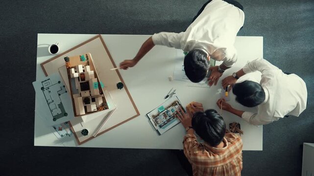 Group of skilled designer looking at model while discussing about building. Top view of architect engineer explain idea while talking and writing blue print scattering at meeting table. Alimentation.