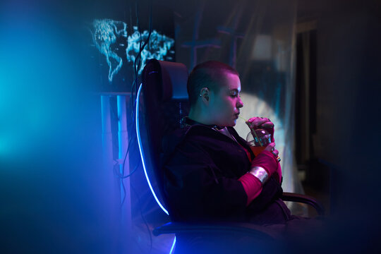 Young adult Caucasian woman sitting in gaming chair holding drink with straw, illuminated by blue light, working on computer with digital world map on screen in dark room - Powered by Adobe
