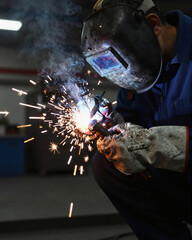 A worker in blue coveralls and a helmet performs arc welding, creating a shower of bright sparks in a dim industrial setting.