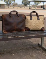 Two contrasting briefcases, one dark leather and one woven straw and leather, rest on a weathered wooden bench in a dry, open outdoor environment.