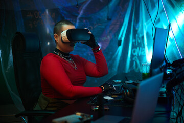 Young adult Caucasian woman with shaved head, wearing virtual reality headset, sitting at computer desk, surrounded by electronic devices engaging in hacking activity in dark room