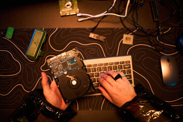 Anonymous young man holding open hard drive while typing on keyboard with other hand, engaging in hacking activity at desk with electronic components and computer mouse visible