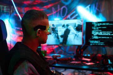 Young adult Caucasian man wearing futuristic glasses, sitting at computer desk, operating multiple monitors with code and surveillance footage, in dark room decorated with graffiti