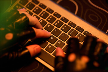 Anonymous young man wearing fingerless gloves typing on laptop keyboard, close up of hands suggesting hacking activity in dark environment, technology concept