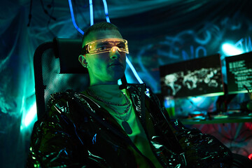 Portrait of young adult Caucasian man, wearing futuristic visor, sitting in dark room illuminated by neon lights, operating multiple computer monitors with digital world map displayed