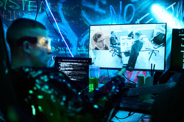 Obraz premium Young adult Caucasian man wearing futuristic glasses sitting at workstation hacking computer system, holding smartphone, monitoring surveillance footage on multiple screens in dark room