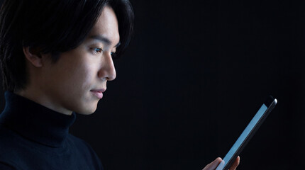 In soft, enchanting illumination, a young Asian man gazes intently at a tablet in a dark room, bathed in the screen's luminous glow.