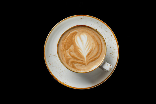 A top-down shot of a cappuccino or latte in a rustic ceramic cup and saucer, featuring delicate latte art on a solid black background