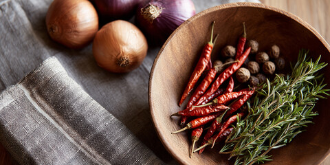 Dried red chili peppers, fresh rosemary, black peppercorns and whole onions arranged in a wooden bowl on natural linen background.