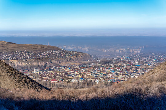 A wide shot from a hill overlooking a Bishkek, the capital of Kyrgyzstan, in a valley, which is heavily covered by a thick layer of atmospheric smog. The air pollution obscures the details of the city