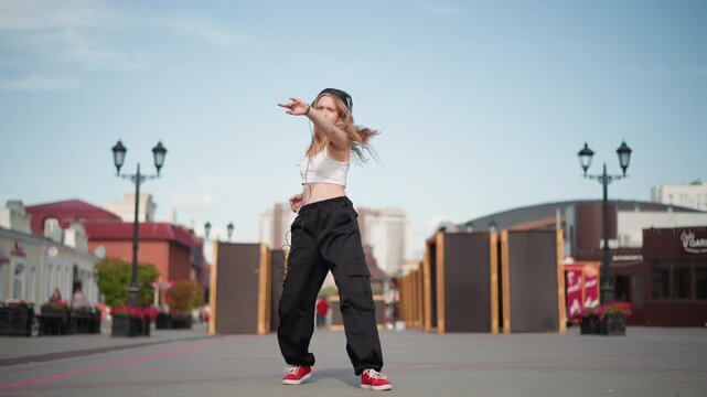 street dancer performing freestyle moves in city square, sunlit empty boulevard with lamp posts and storefronts, loose black cargo pants and red sneakers, black beanie, expressive arm gestures