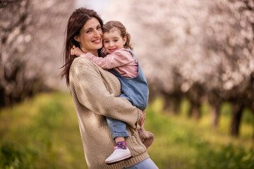 Fototapeta premium Mother and daughter smiling while embracing warmly, experiencing a joyful family moment in a beautiful outdoor almond tree field with white blossoms in spring