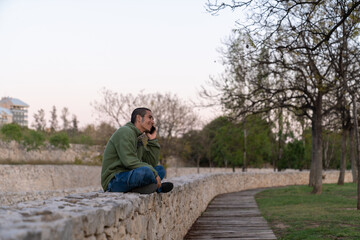 A captivating dusk silhouette captures a young brunette man in a green hoodie and jeans sitting cross-legged on a white stone wall, engaged in an intriguing phone conversation.  © Araguatai