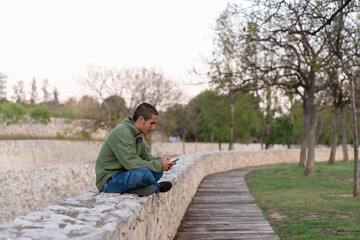A striking dusk silhouette captures a young brunette man in green hoodie and jeans sitting cross-legged on a white stone wall, intently focused on his smartphone © Araguatai