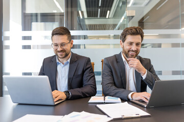Two smiling professional businessmen are working diligently on their laptops at an office desk, illustrating modern communication, teamwork, and business technology in a corporate setting