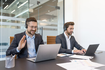 Two male professionals wearing headsets and suits engaging in a video call and typing on laptops, showing teamwork and communication within a corporate customer support environment