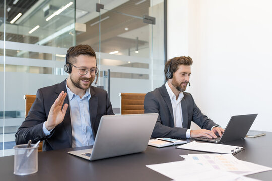 Two male professionals wearing headsets and suits engaging in a video call and typing on laptops, showing teamwork and communication within a corporate customer support environment - Powered by Adobe