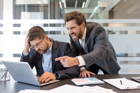 Stressed employee with head in hand experiences workplace harassment from shouting, angry boss pointing and criticizing his work on a laptop, causing conflict and frustration in the office