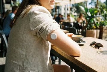Woman wearing a health monitor on her arm while sitting at an outdoor cafe table with coffee and sunglasses