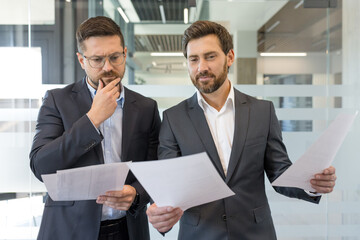Two professional businessmen are collaborating and carefully analyzing financial documents and paperwork, working together to make important business decisions in a modern office environment