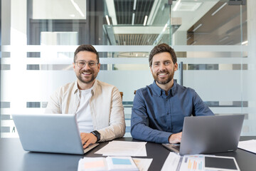 Two smiling male colleagues collaborate at a bright coworking desk, laptops open and paperwork spread as they plan a startup project, discussing strategy and ideas