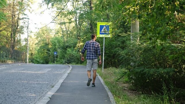 A man walks on peaceful sidewalk surrounded by lush summer greenery, moving calmly along the road edge. The concept emphasizes routine, movement, lifestyle, and everyday outdoor activity. Rear view