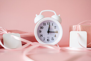 White alarm clock with pink gift boxes and blank gift tags on pink background. Time for celebration, Valentine's Day or Birthday concept