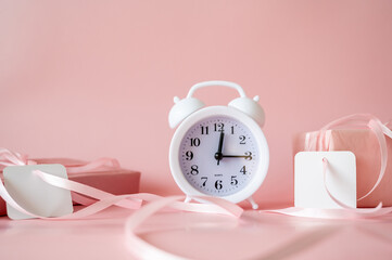 White alarm clock with pink gift boxes and blank gift tags on pink background. Time for celebration, Valentine's Day or Birthday concept
