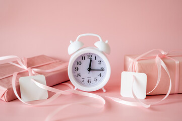 White alarm clock with pink gift boxes and blank gift tags on pink background. Time for celebration, Valentine's Day or Birthday concept