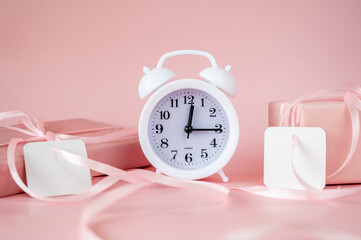 White alarm clock with pink gift boxes and blank gift tags on pink background. Time for celebration, Valentine's Day or Birthday concept