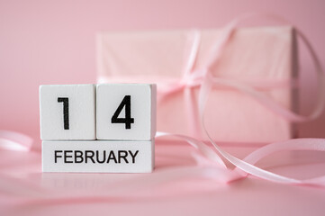 February 14 wooden block calendar on pink background with blurred gift box and ribbons. Valentine's Day celebration concept