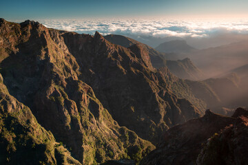 Obraz premium Rugged peaks and deep valleys of Madeira’s highest mountains glow in warm morning sunlight above a thick sea of clouds, seen from Pedra Rija viewpoint under a clear blue sky